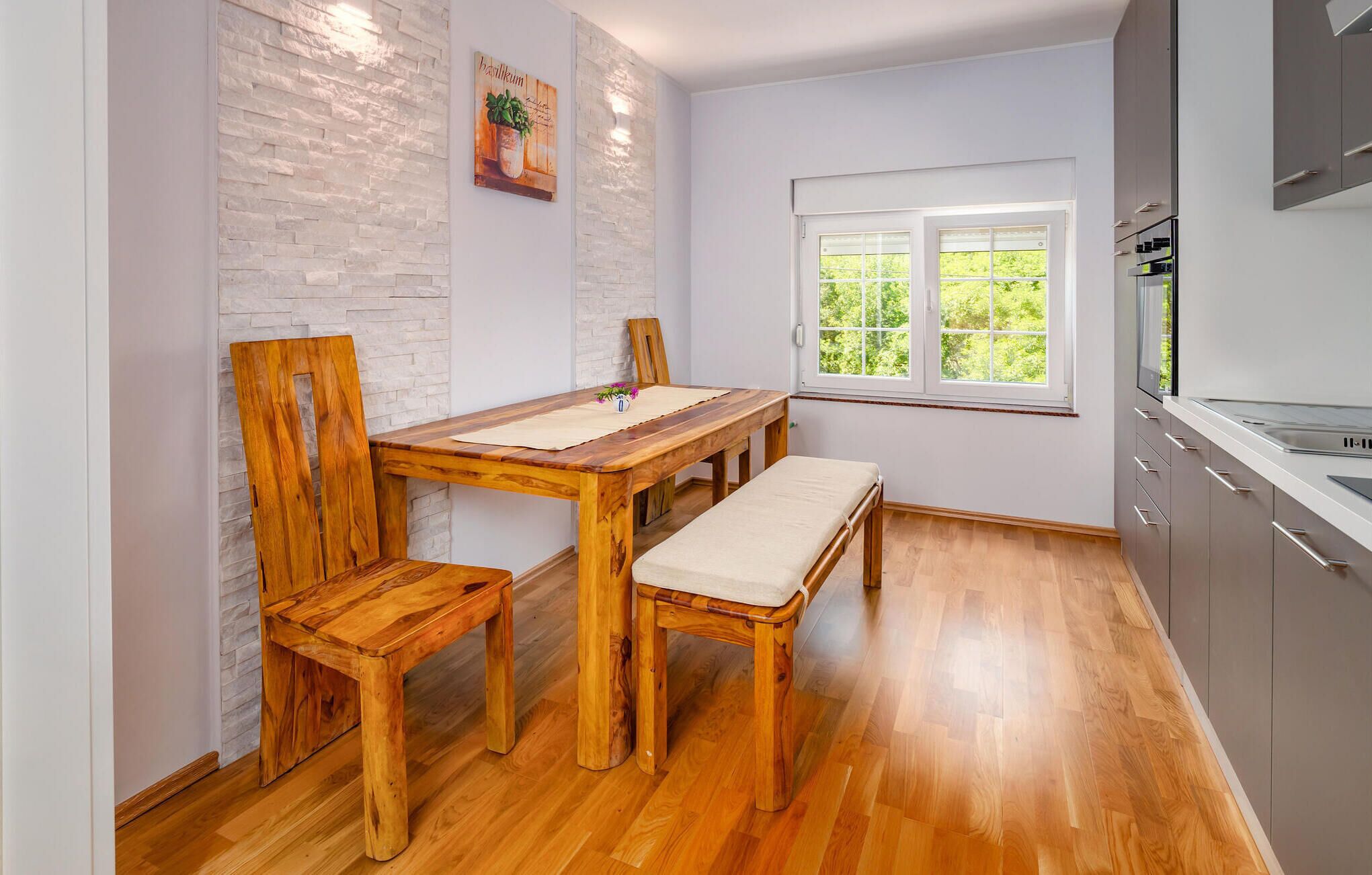 A small kitchen with a wooden table and chairs, a window with a view of greenery, and a modern stove.