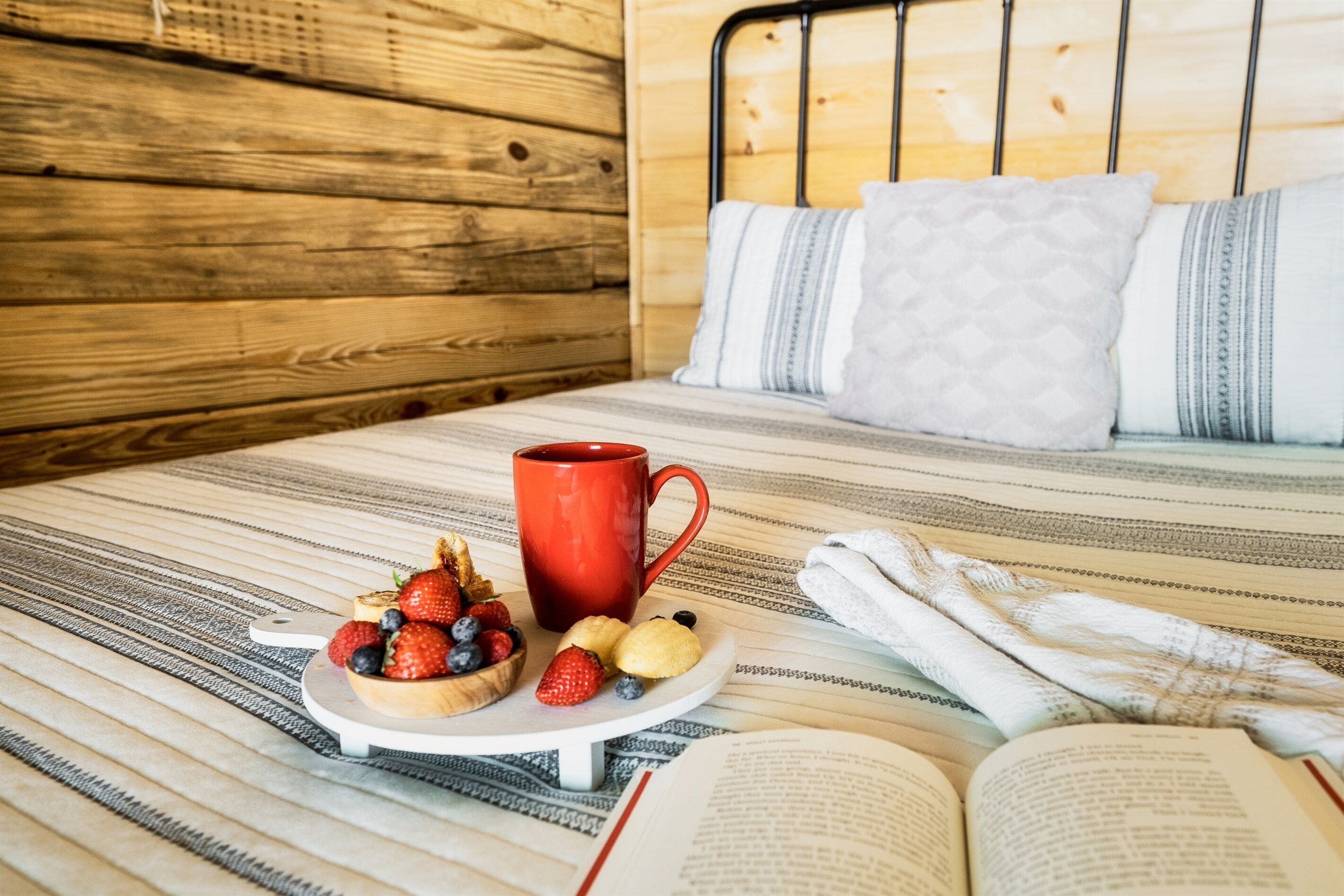 Una cama con cabecera de madera, una colcha a rayas y una taza roja sobre una mesita auxiliar.