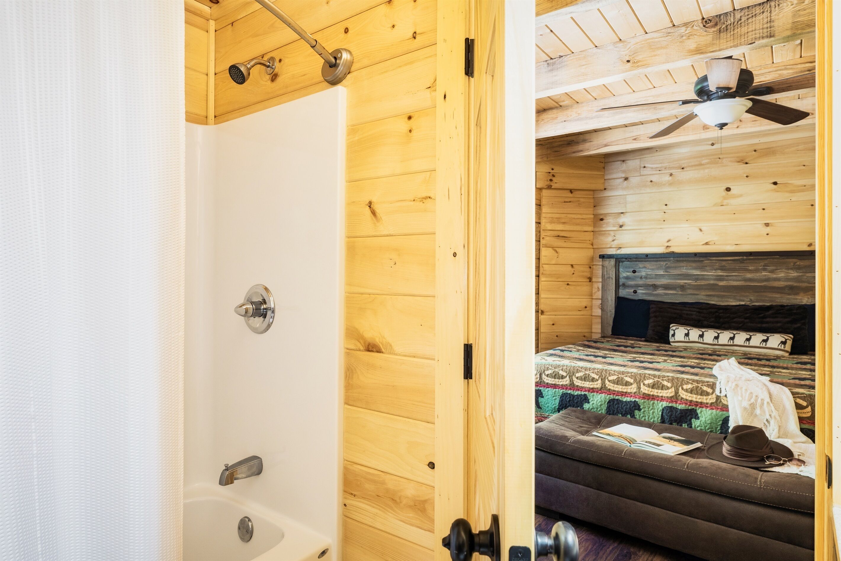 Ein Holzhütten-Zimmer mit Dusche, einem Bett mit gemustertem Bettbezug und einem Deckenventilator.