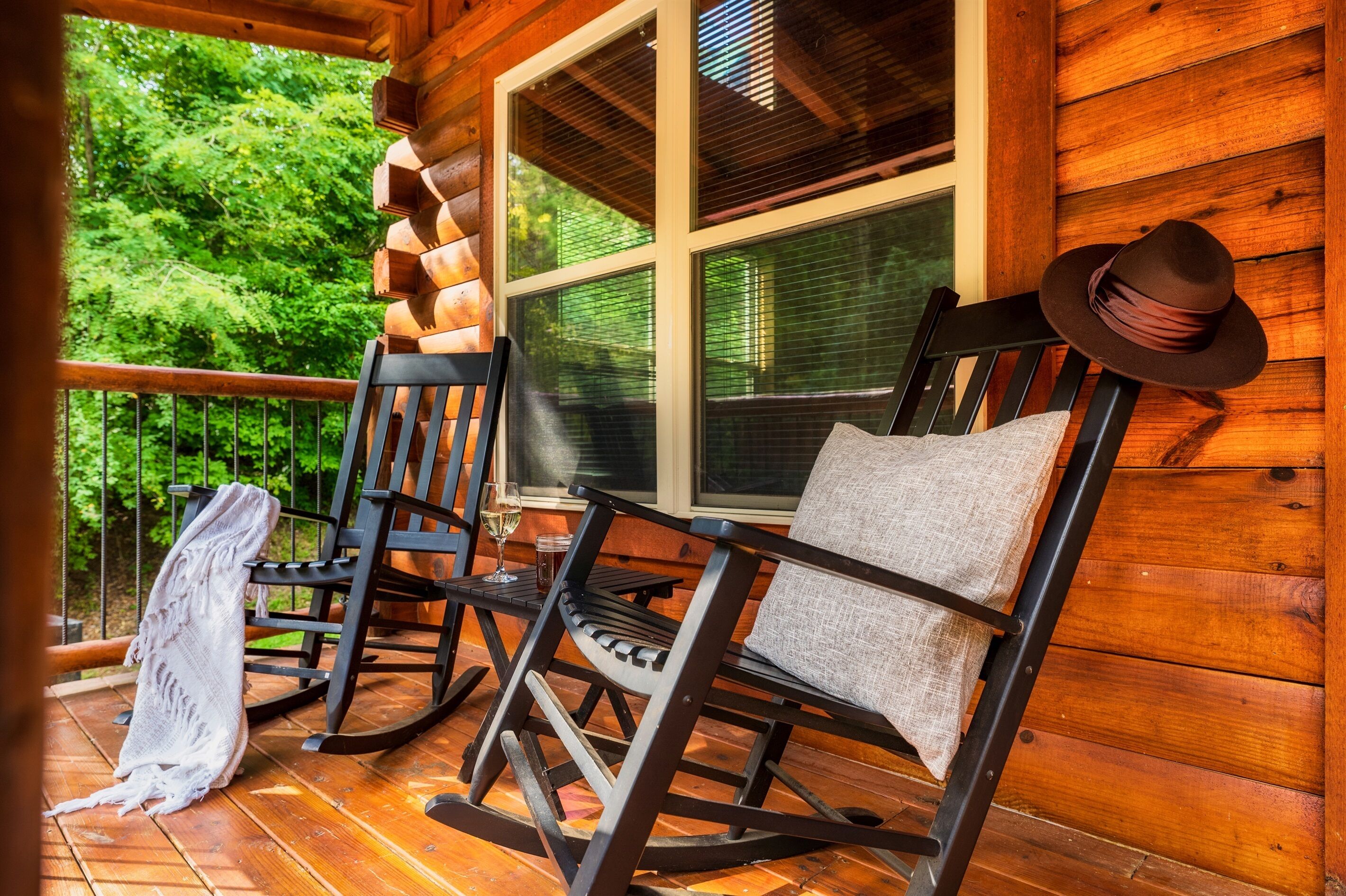 A wooden porch with rocking chairs and a bench, a window with blinds, and a view of greenery.