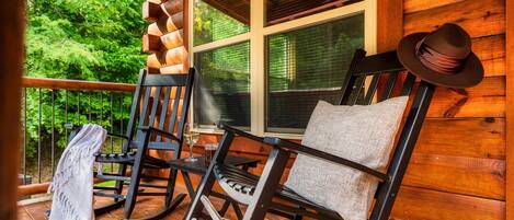 A wooden porch with rocking chairs and a bench, a window with blinds, and a view of greenery.