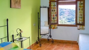 A room with a bed, a small table with a lamp, a metal stand with a mirror, and a window with curtains.