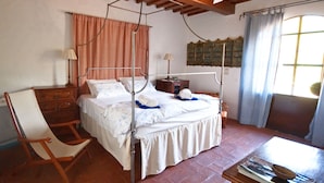 Een slaapkamer met een hemelbed, een houten stoel, een nachtkastje en een raam met gordijnen.