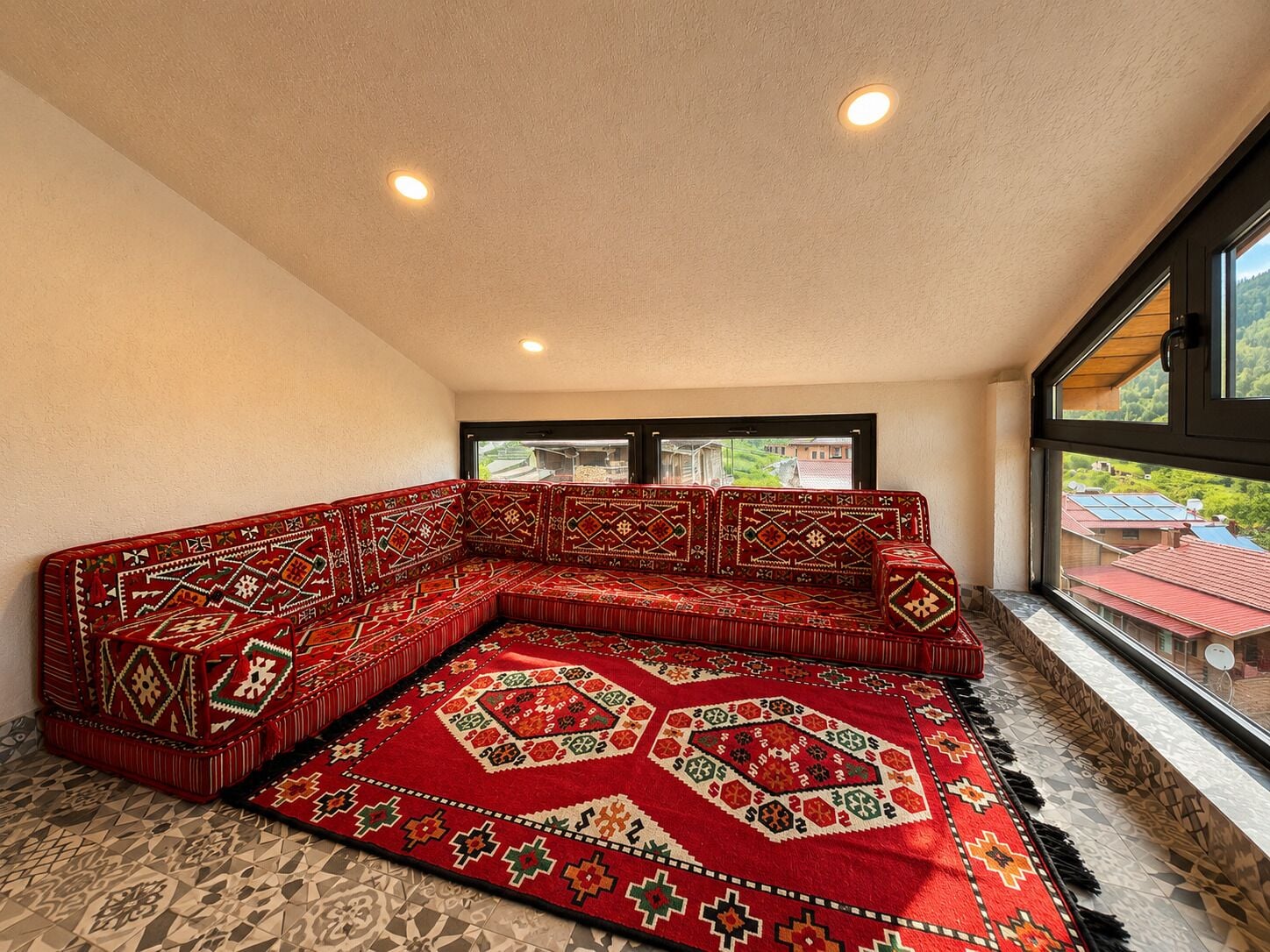 A corner room with a large red patterned sofa, a window with a view of houses, and a ceiling with recessed lighting.