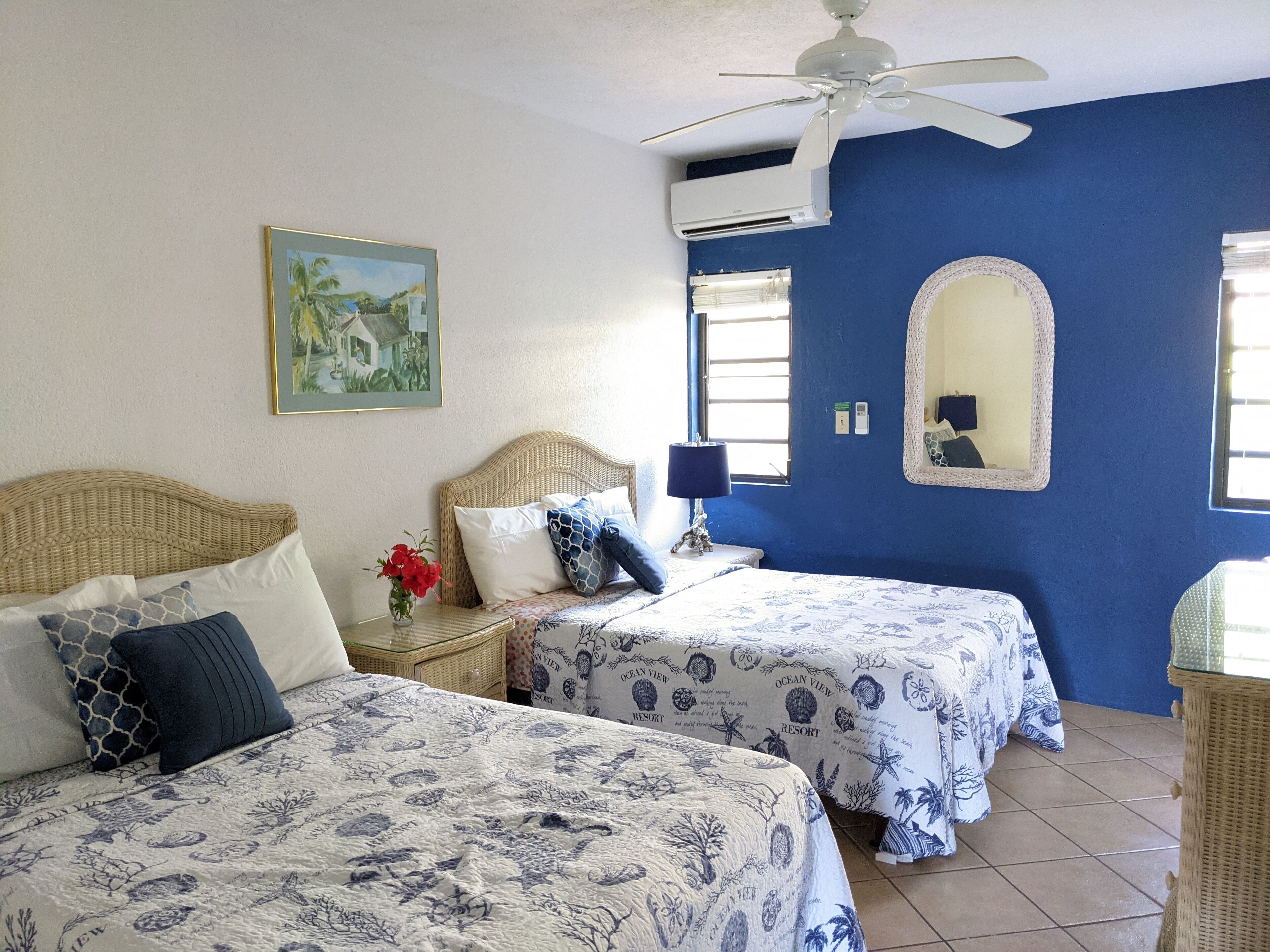 Habitación con dos camas, ventilador de techo, equipo de aire acondicionado, un cuadro enmarcado, un espejo y una ventana con persianas.