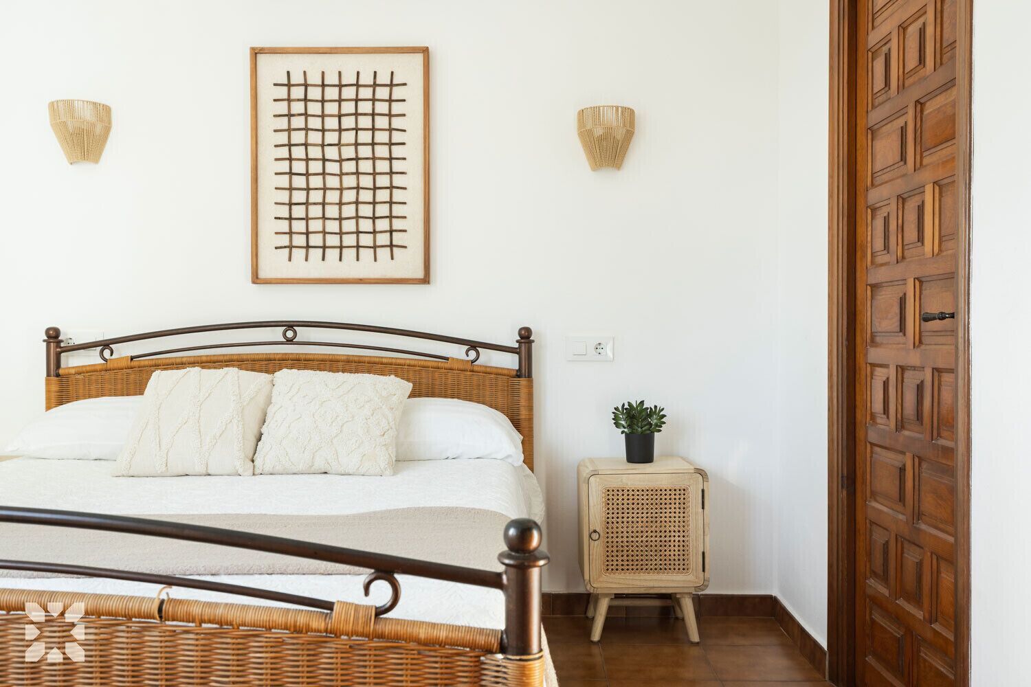 Una cama con cabecera de madera, una mesita de mimbre con una planta y una puerta de madera.