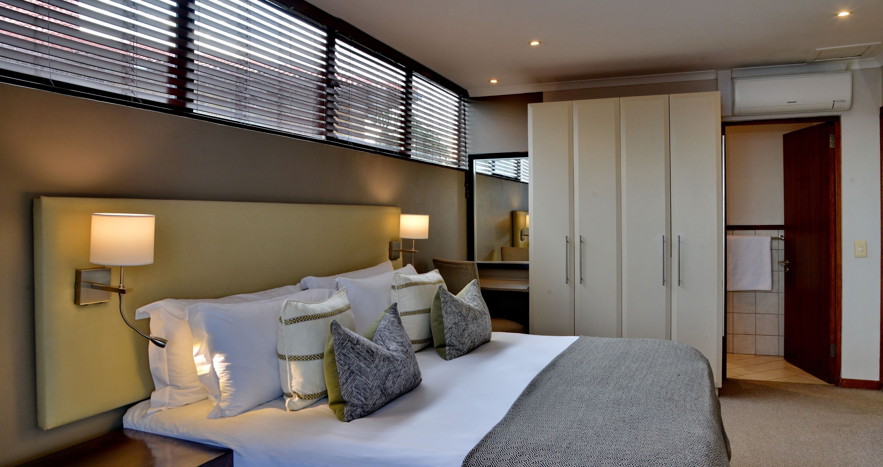 Una habitación de hotel moderna con una cama grande, lámparas de noche, un armario y un baño visible a través de una puerta abierta.