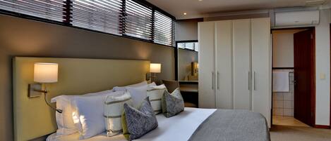 Una habitación de hotel moderna con una cama grande, lámparas de noche, un armario y un baño visible a través de una puerta abierta.