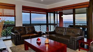 Una sala de estar moderna con sofás de cuero, una mesa de centro de madera y una estructura de vidrio con una vela. Amplias ventanas ofrecen vistas al océano y un balcón con barandilla.