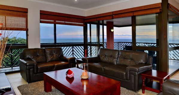 Una sala de estar moderna con sofás de cuero, una mesa de centro de madera y una estructura de vidrio con una vela. Amplias ventanas ofrecen vistas al océano y un balcón con barandilla.