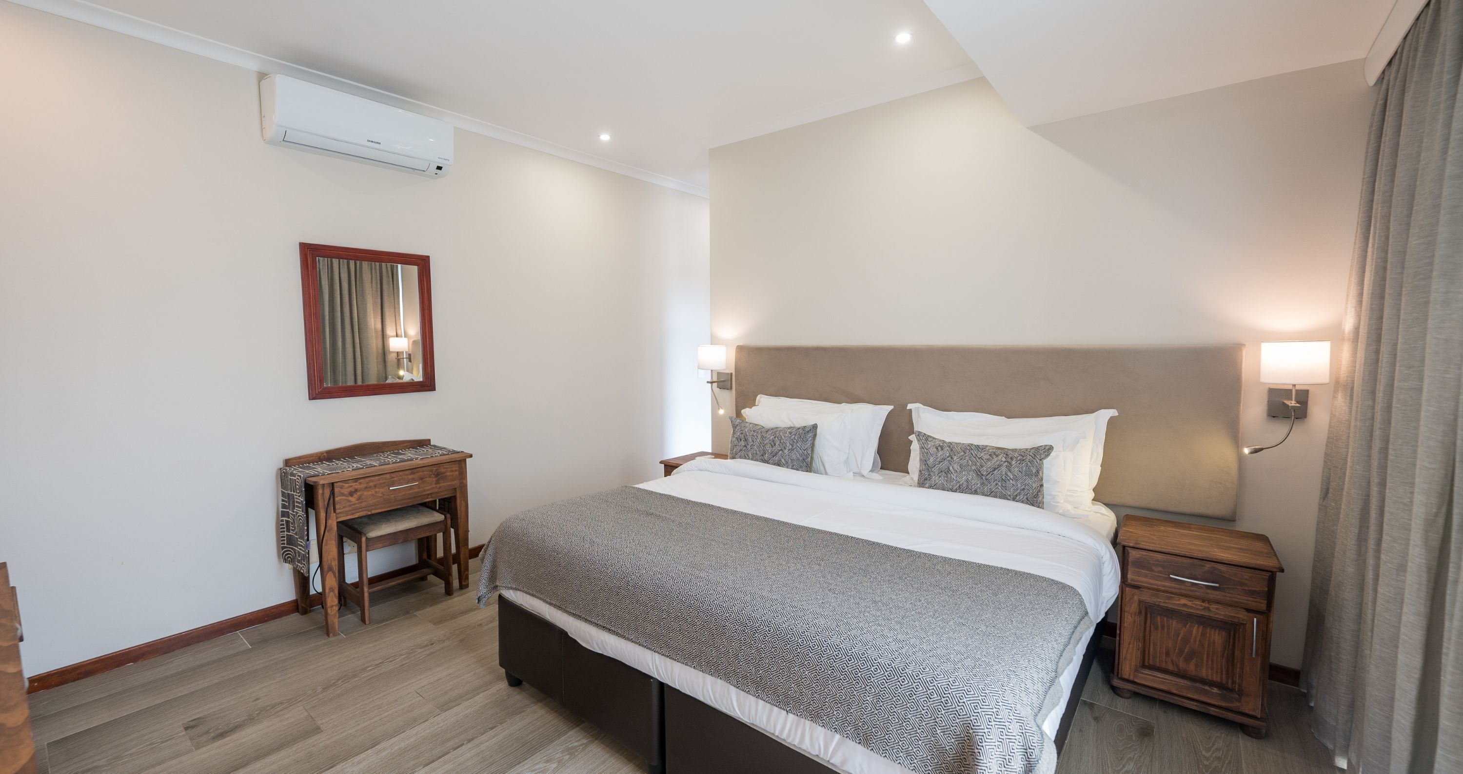 A hotel room with a large bed, bedside tables, a desk, and an air conditioning unit.