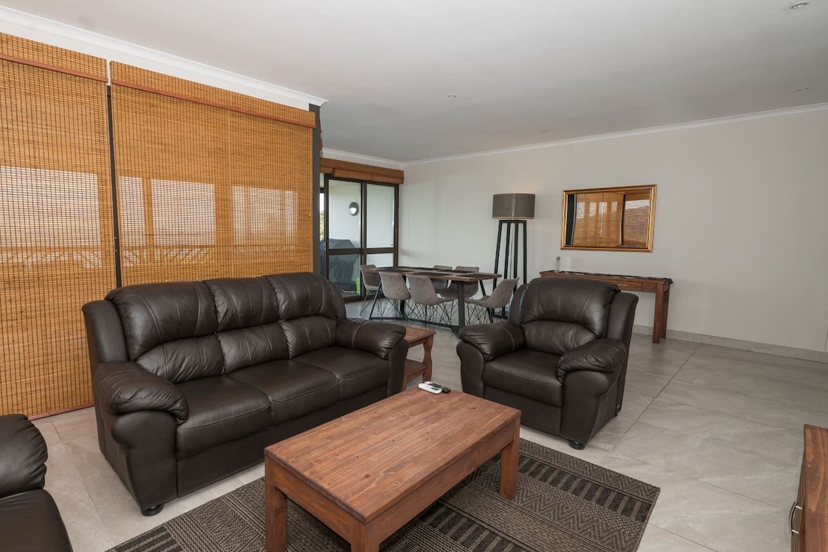 Ein Wohnzimmer mit Sofa, Sessel und Couchtisch. Im Hintergrund ist eine Essecke mit Tisch und Stühlen zu sehen.