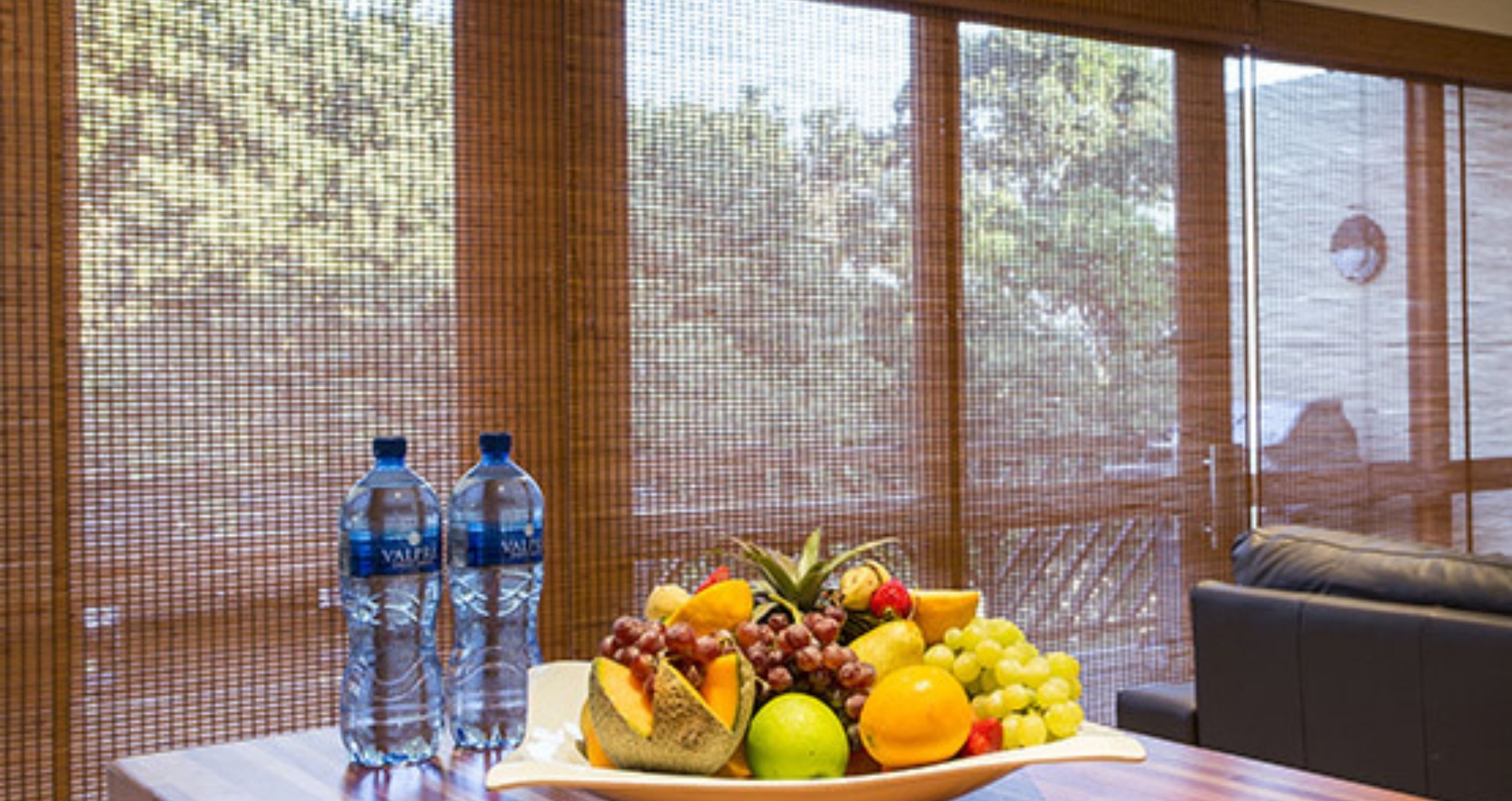 Una mesa con una bandeja de frutas y dos botellas de agua frente a una ventana con vista a árboles.