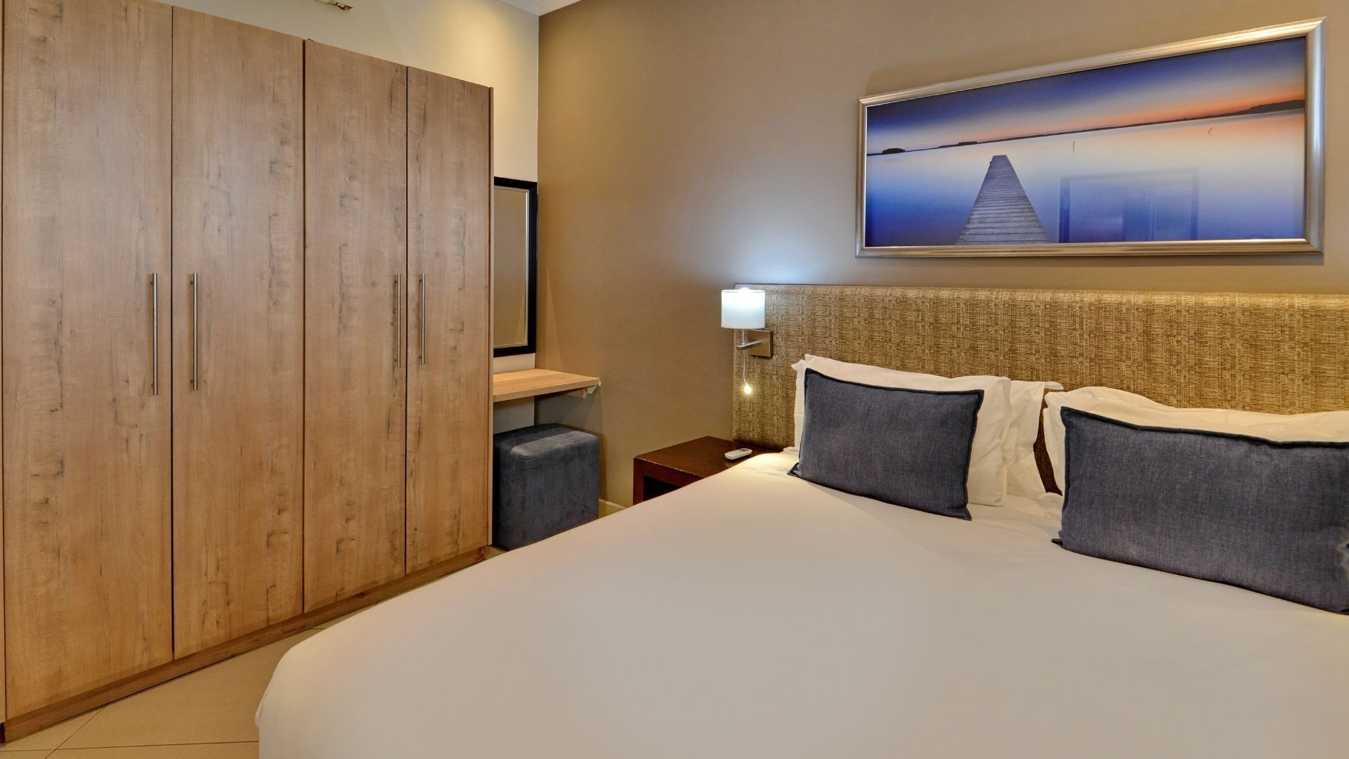 Una habitación de hotel con un armario de madera grande, una cama con dos almohadas, una mesita de noche, una lámpara y un cuadro de un atardecer sobre un muelle.
