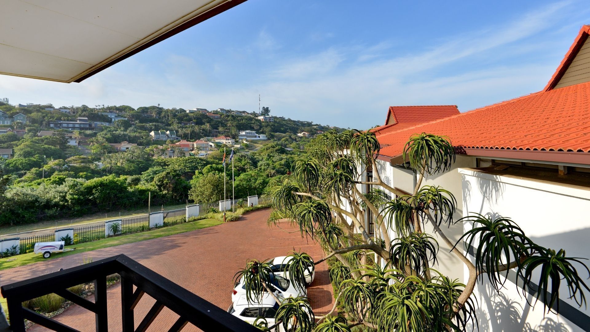 A balcony with a view of a residential area, a parked car, and a red-roofed building.