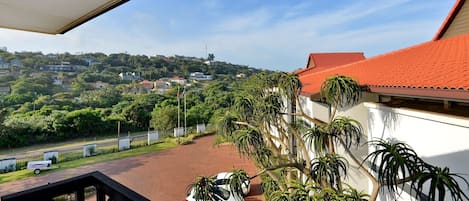 A balcony with a view of a residential area, a parked car, and a red-roofed building.