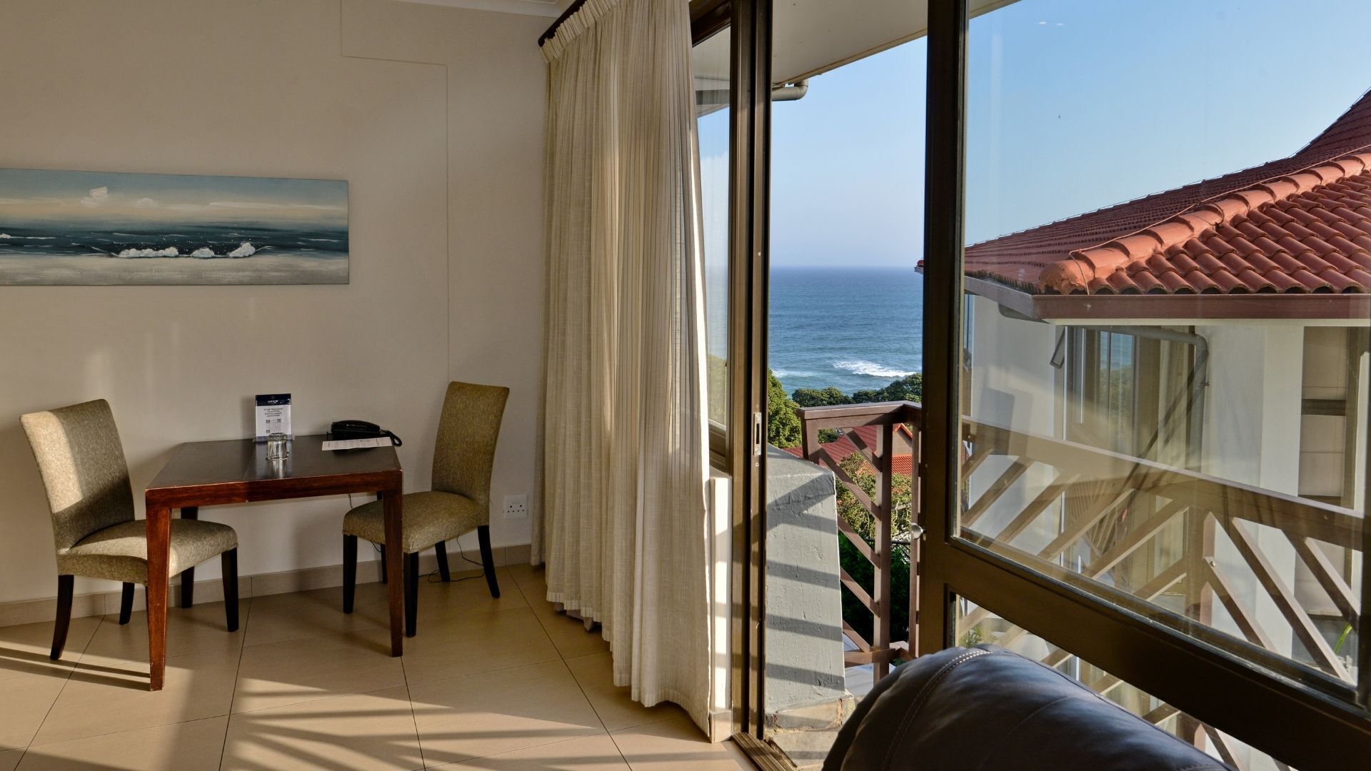 A room with a view of the ocean, a table with chairs, and a window with curtains.