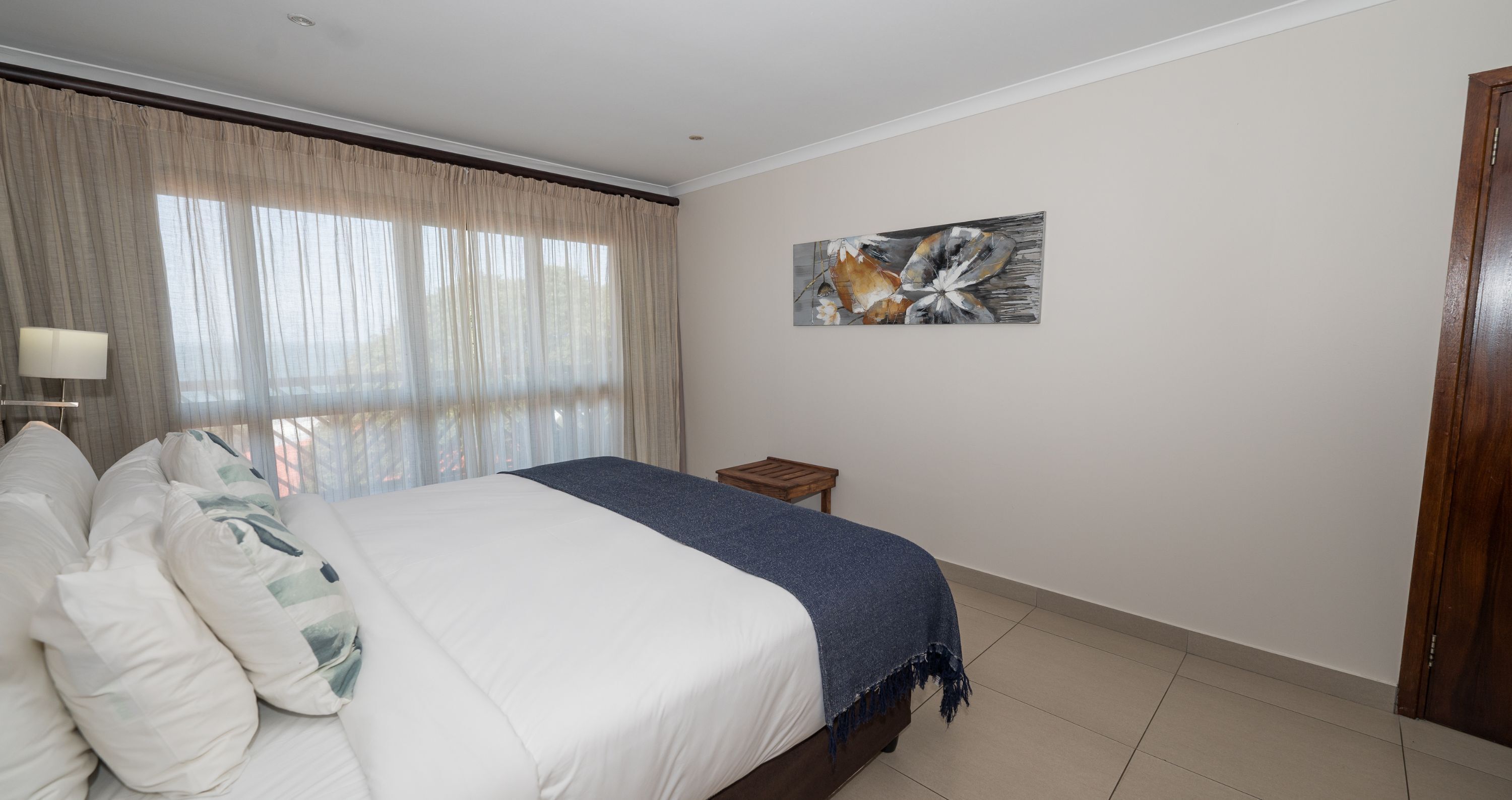 Un dormitorio con cama, mesita de noche, lámpara, ventana con cortinas y un cuadro en la pared.