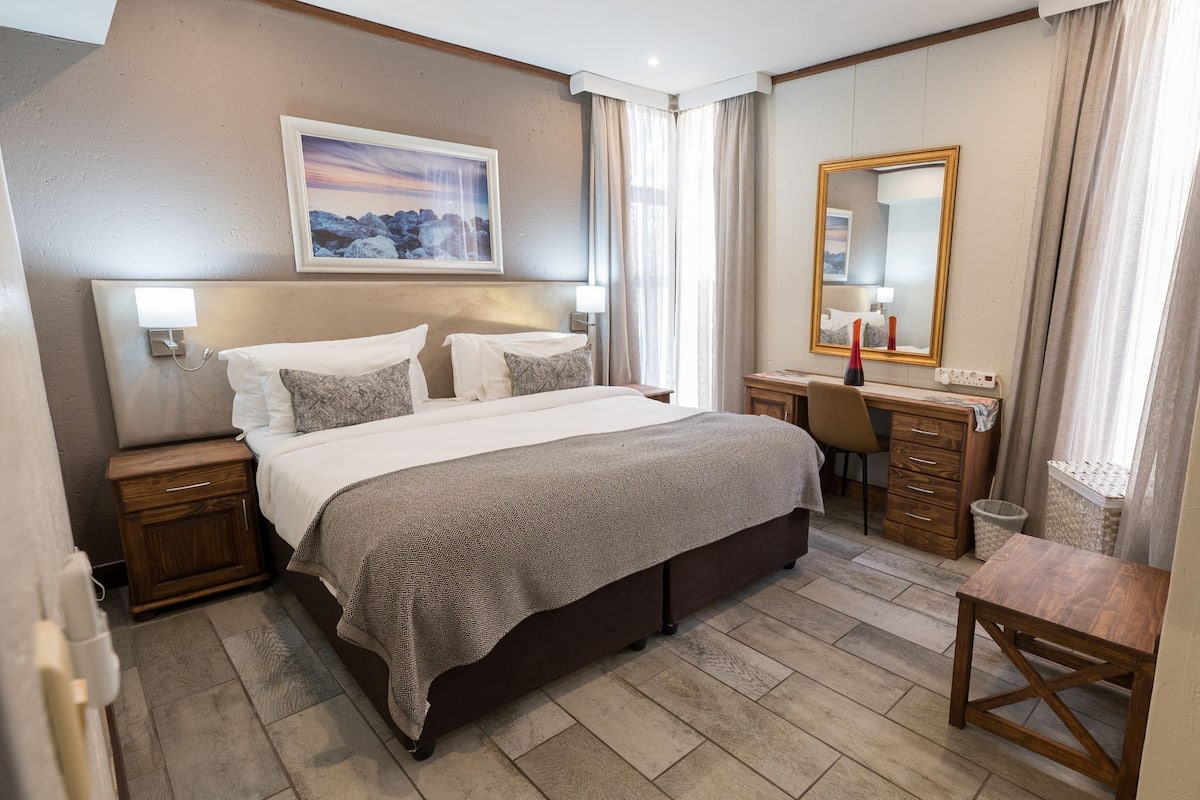 Ein Hotelzimmer mit Bett, Nachttischen, Schreibtisch, Stuhl, Spiegel und einem Bild an der Wand.
