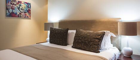 A neatly made bed with a brown bedspread and two decorative pillows, a wall art piece, and a bedside lamp on a wooden nightstand.