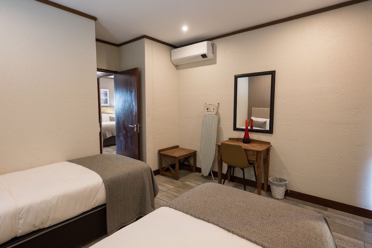 Ein Hotelzimmer mit zwei Einzelbetten, einem kleinen Schreibtisch mit Stuhl, einem Spiegel und einem Mülleimer.