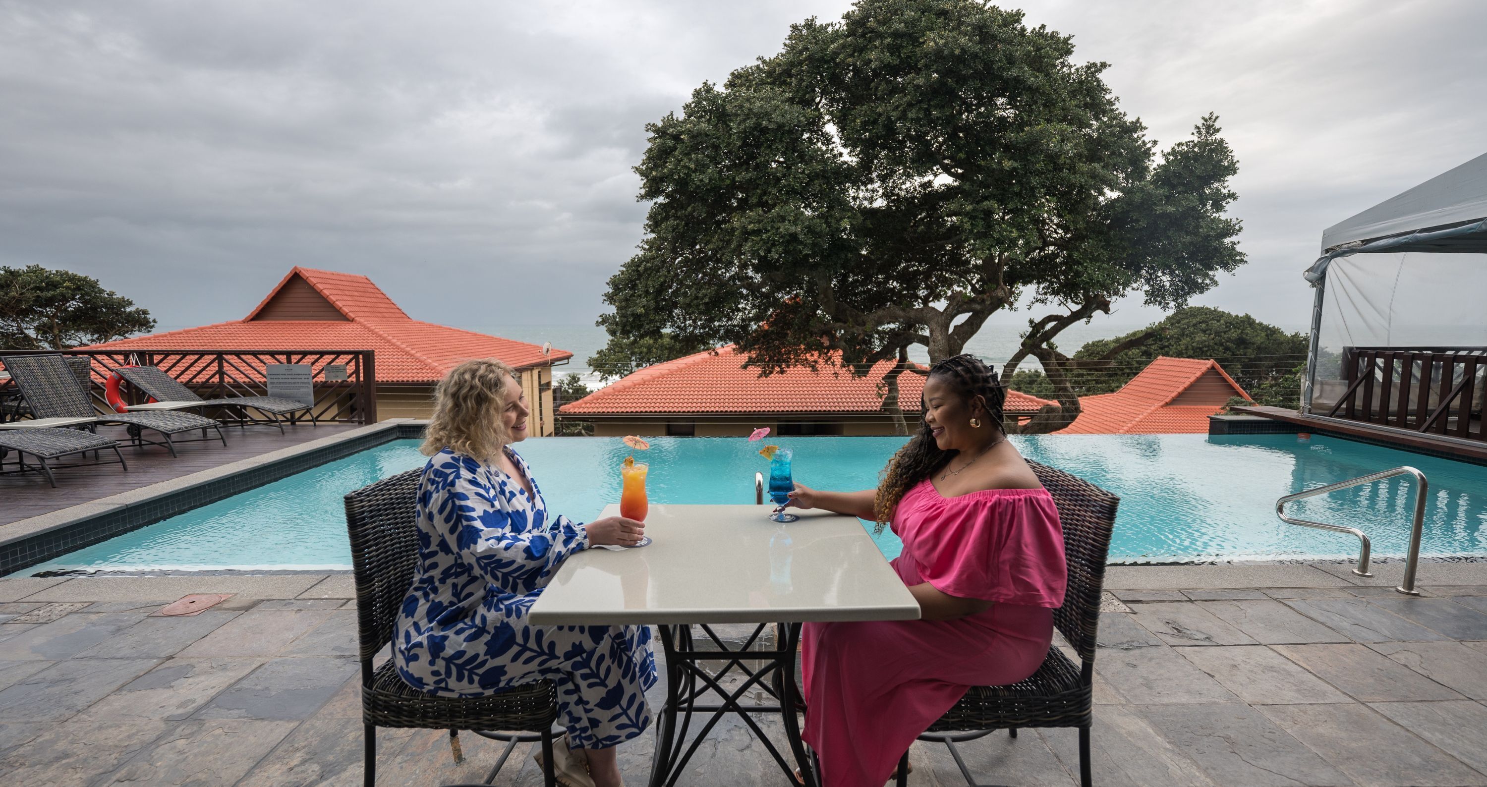 Two people seated at a table by a pool with a view of a building with red roofs and a large tree in the background.