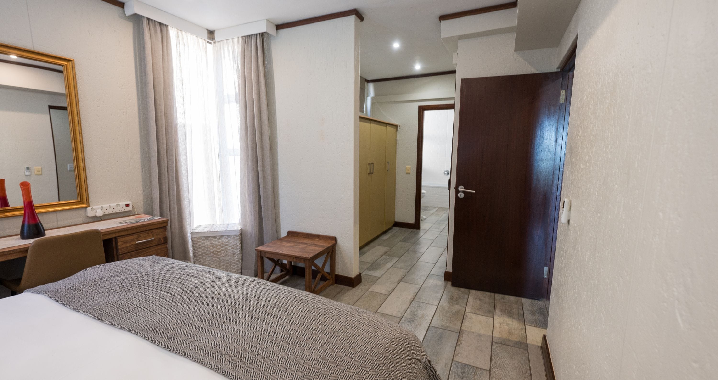 Een hotelkamer met een bed, een bureau, een stoel, een tafeltje, een spiegel, een kledingkast en een deur.