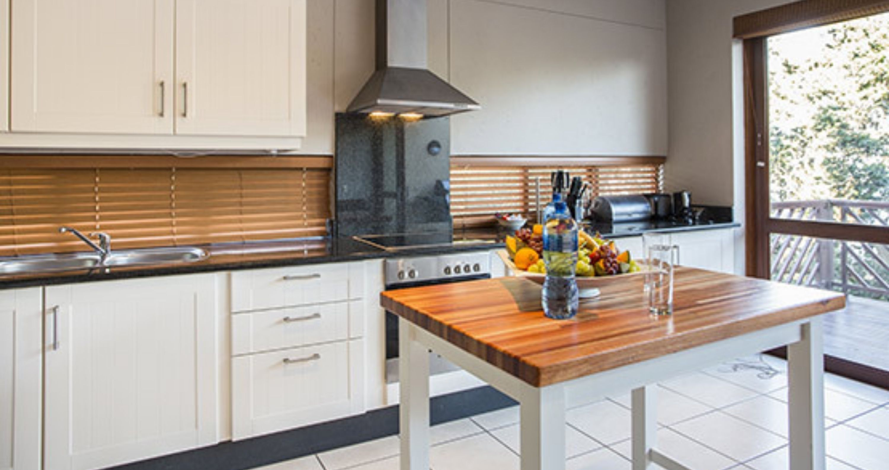 A modern kitchen with white cabinets, a stainless steel sink, a gas stove, and a wooden table with a fruit bowl.