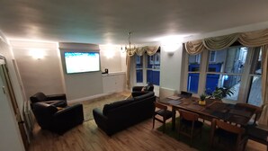 Un salón con suelo de madera, un sofá negro, una mesa de comedor con sillas, un televisor en la pared y una ventana con cortinas.