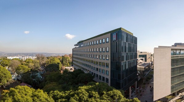 Un edificio moderno a più piani con facciata in vetro, circondato da vegetazione lussureggiante e altri edifici sullo sfondo.