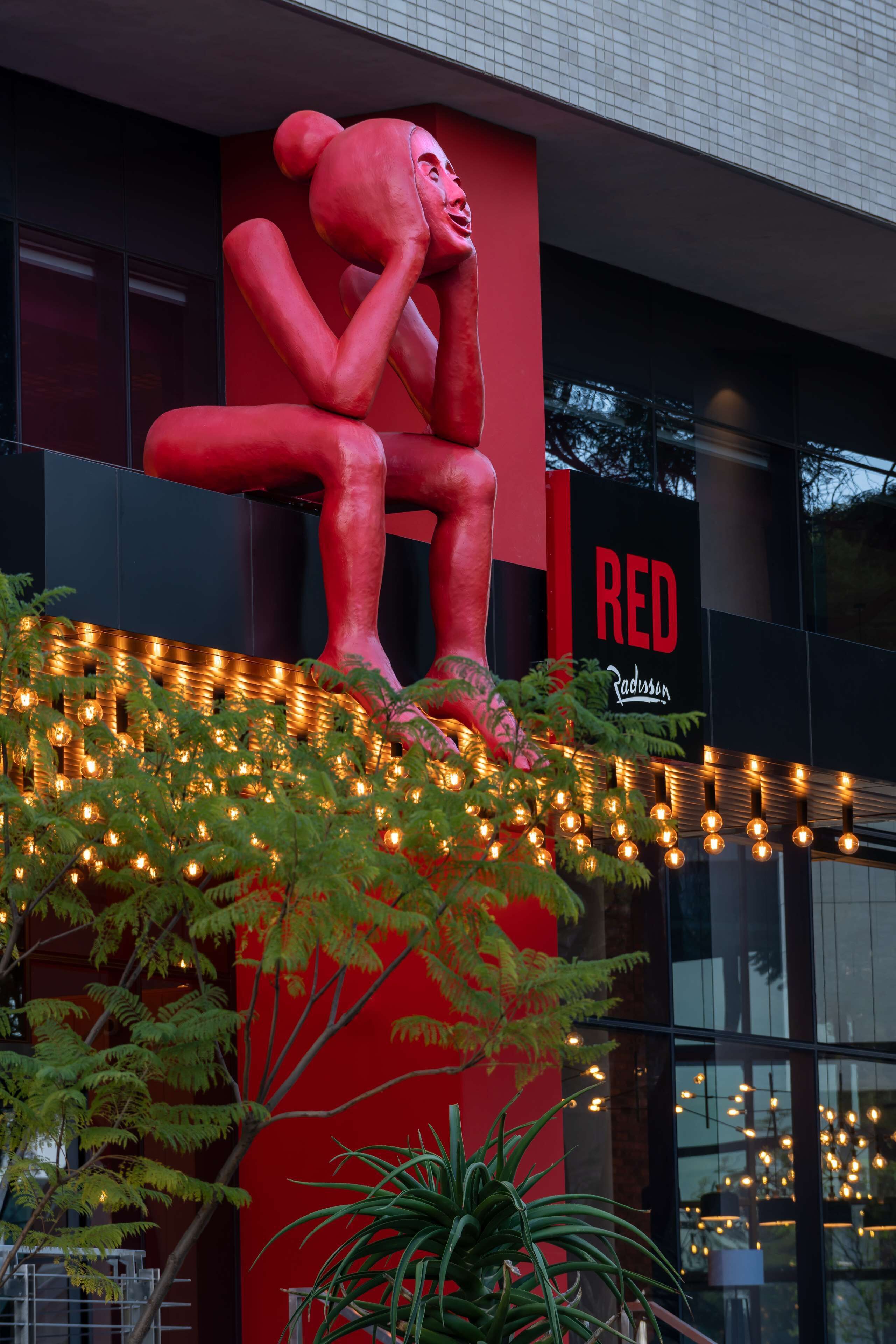 A building with a large red sculpture of a seated figure, a sign reading "RED Radisson", and string lights on the ground.