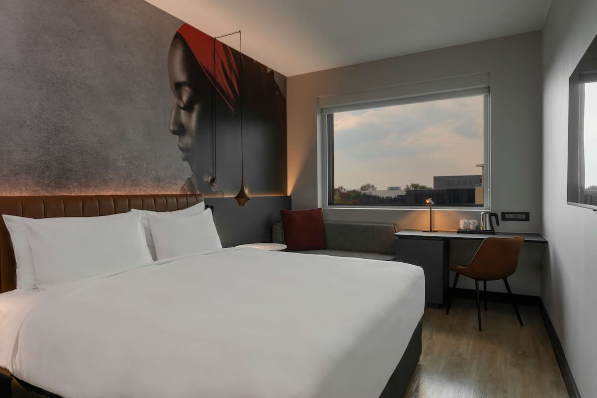 Habitación de hotel con una cama grande, un escritorio con silla, una ventana con vistas a la ciudad y un cuadro de gran tamaño en la pared.