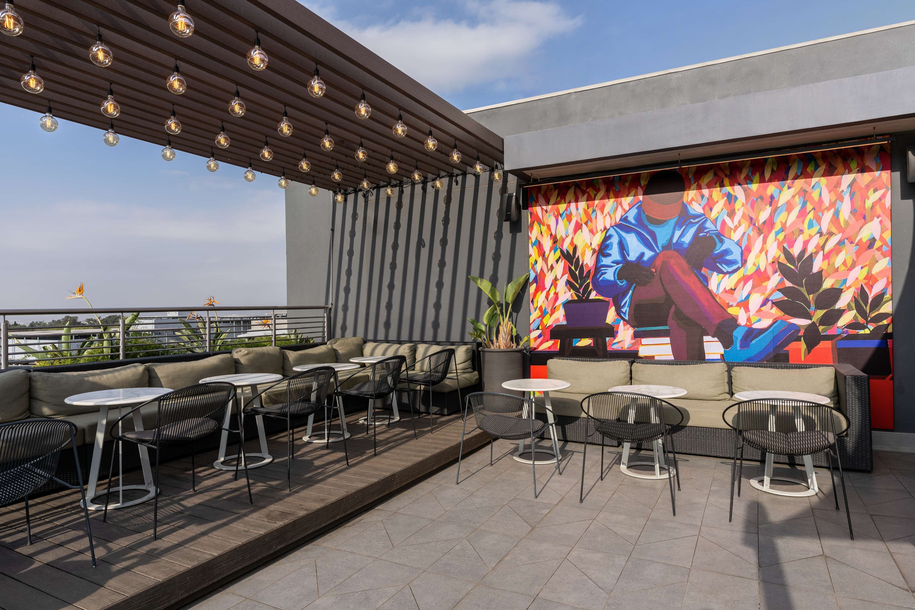 Un espacio al aire libre con mesas y sillas, un mural y una p érgola con luces colgantes.
