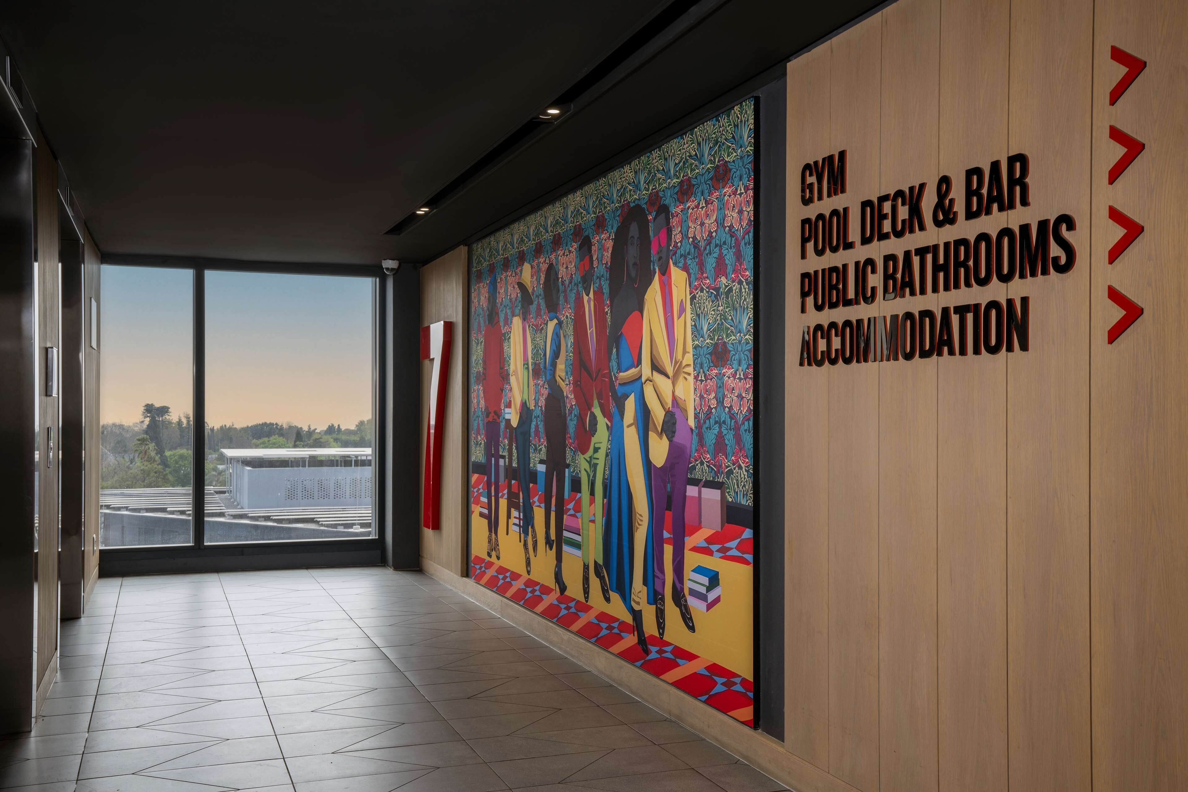 Un pasillo con un gran mural, vistas a través de puertas de cristal y un letrero que indica servicios como gimnasio, terraza de la piscina, bar, baños públicos y alojamiento.