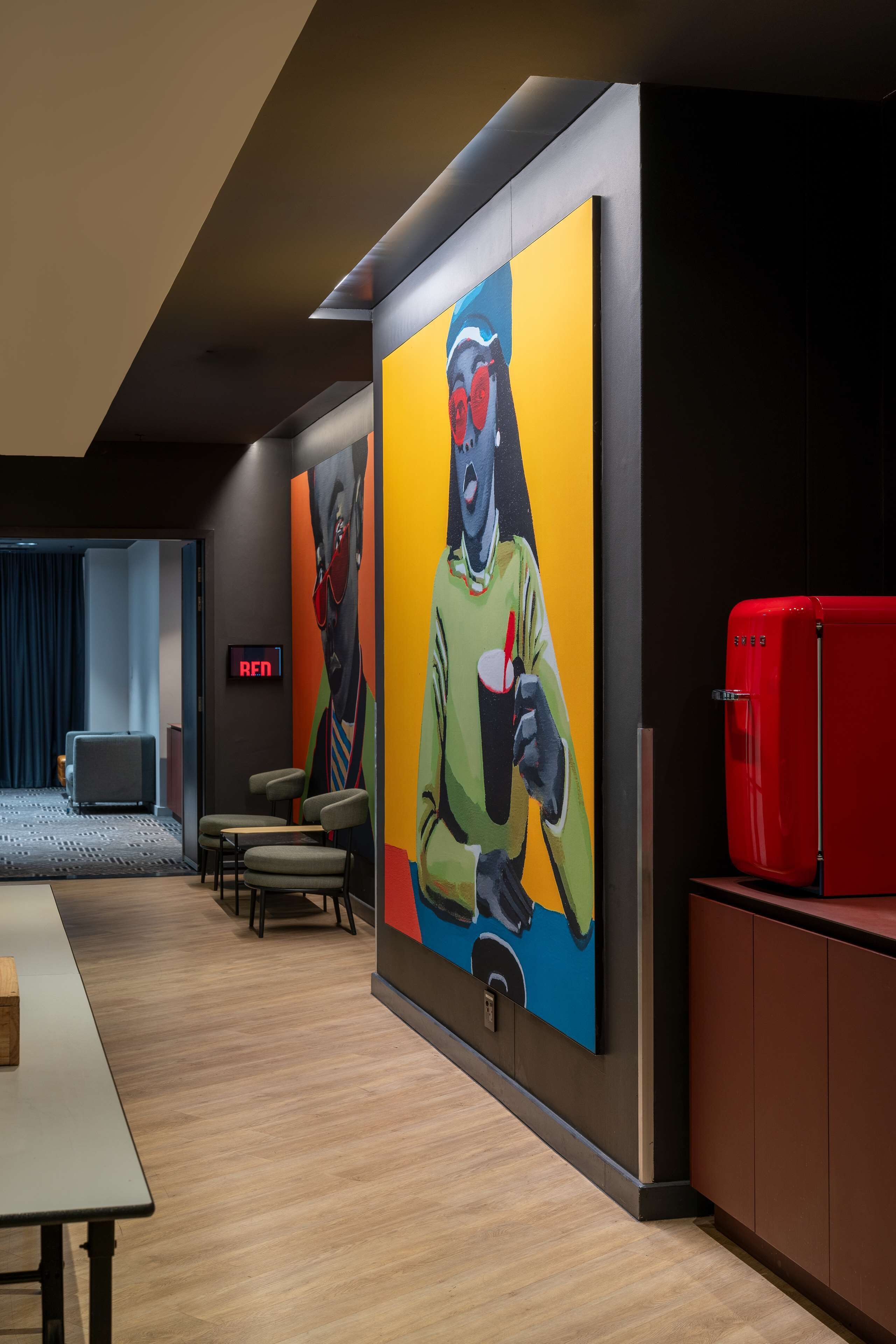 Eine moderne Hotellobby mit einer großen, farbenfrohen Wandmalerei, einem roten Kühlschrank und einer Sitzecke mit Tischen und Stühlen.