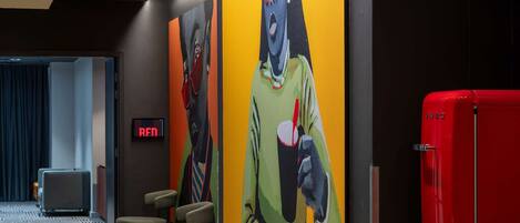 Eine moderne Hotellobby mit einer großen, farbenfrohen Wandmalerei, einem roten Kühlschrank und einer Sitzecke mit Tischen und Stühlen.