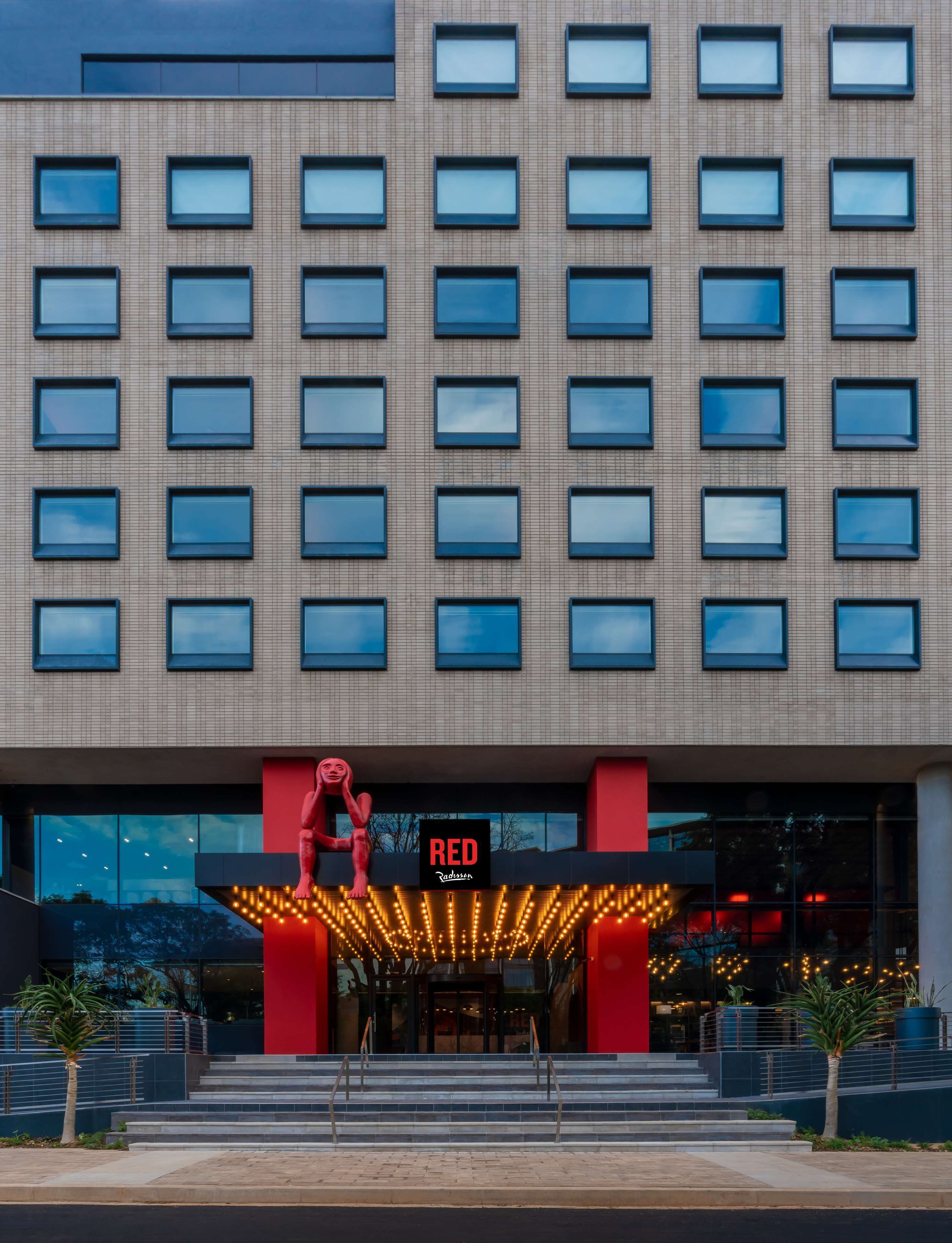 A modern hotel facade with a grid of windows and a prominent entrance featuring a red sculpture and illuminated signage.