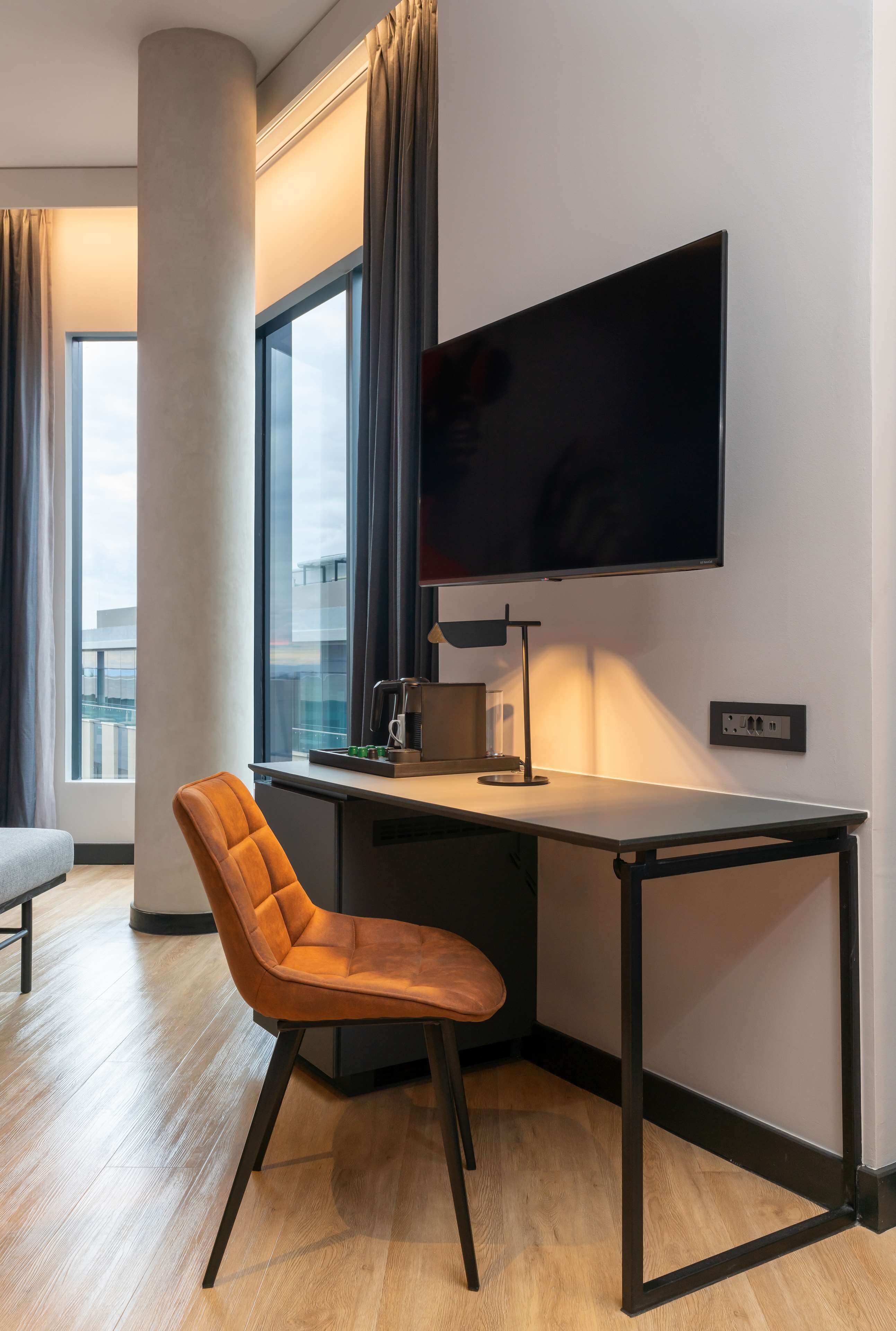 Una habitación de hotel moderna con un ventanal grande, un televisor de pantalla plana en la pared, un escritorio con lámpara y una silla cómoda.