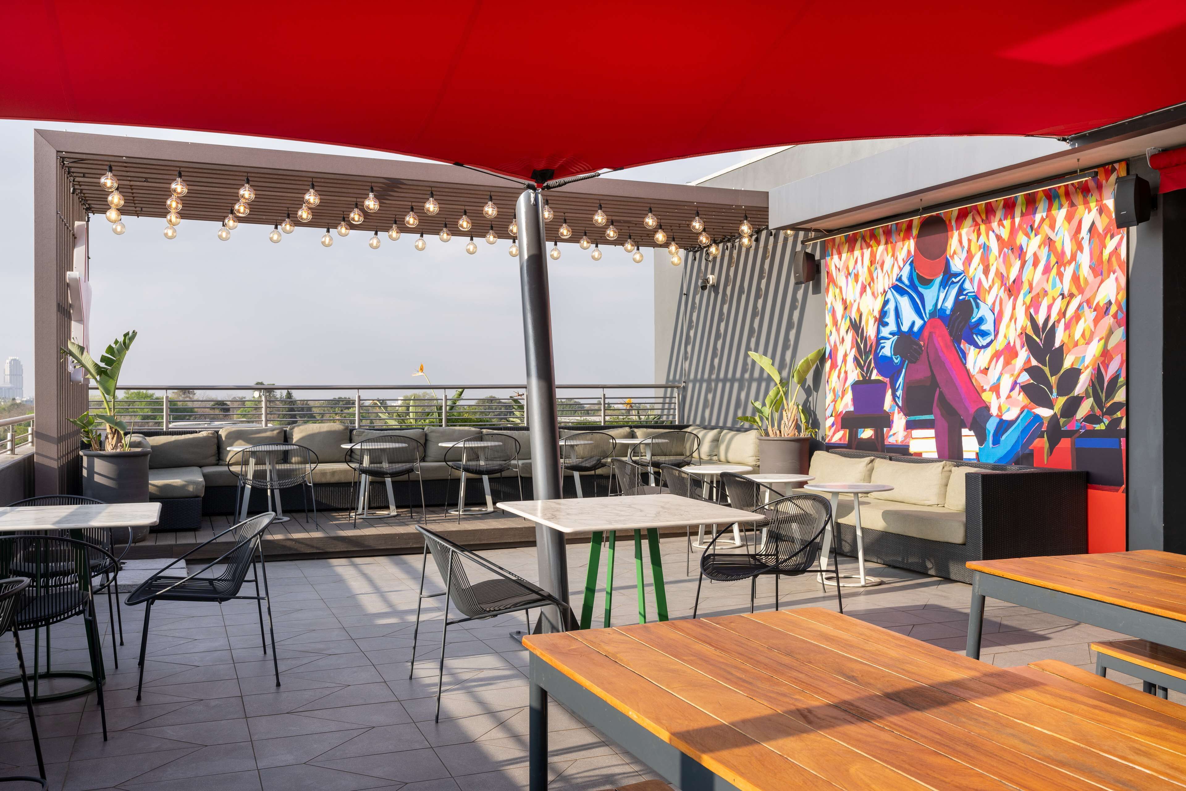 Un espacio al aire libre con mesas y sillas, un mural en la pared y vistas de la ciudad.