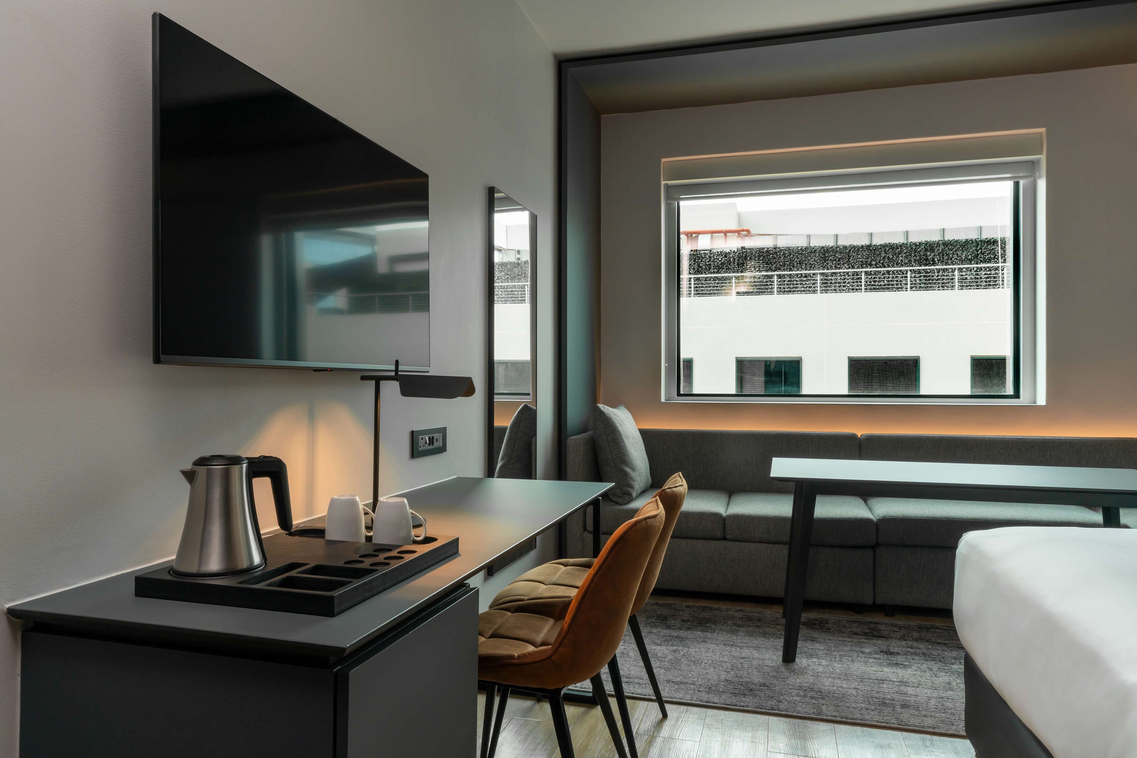 A modern hotel room with a flat-screen TV, a small desk with a kettle, a sofa, and a large window offering a city view.