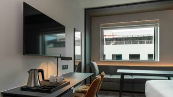 A modern hotel room with a flat-screen TV, a small desk with a kettle, a sofa, and a large window offering a city view.