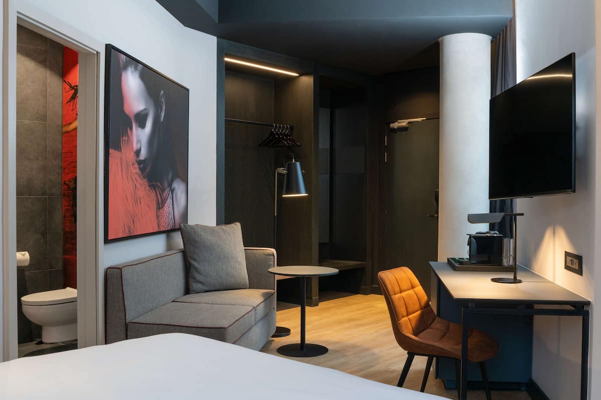 Una habitación de hotel moderna con una cama, un sofá gris, una mesita redonda pequeña, un sillón naranja, un escritorio con lámpara, un televisor y una obra de arte grande con marco en la pared.