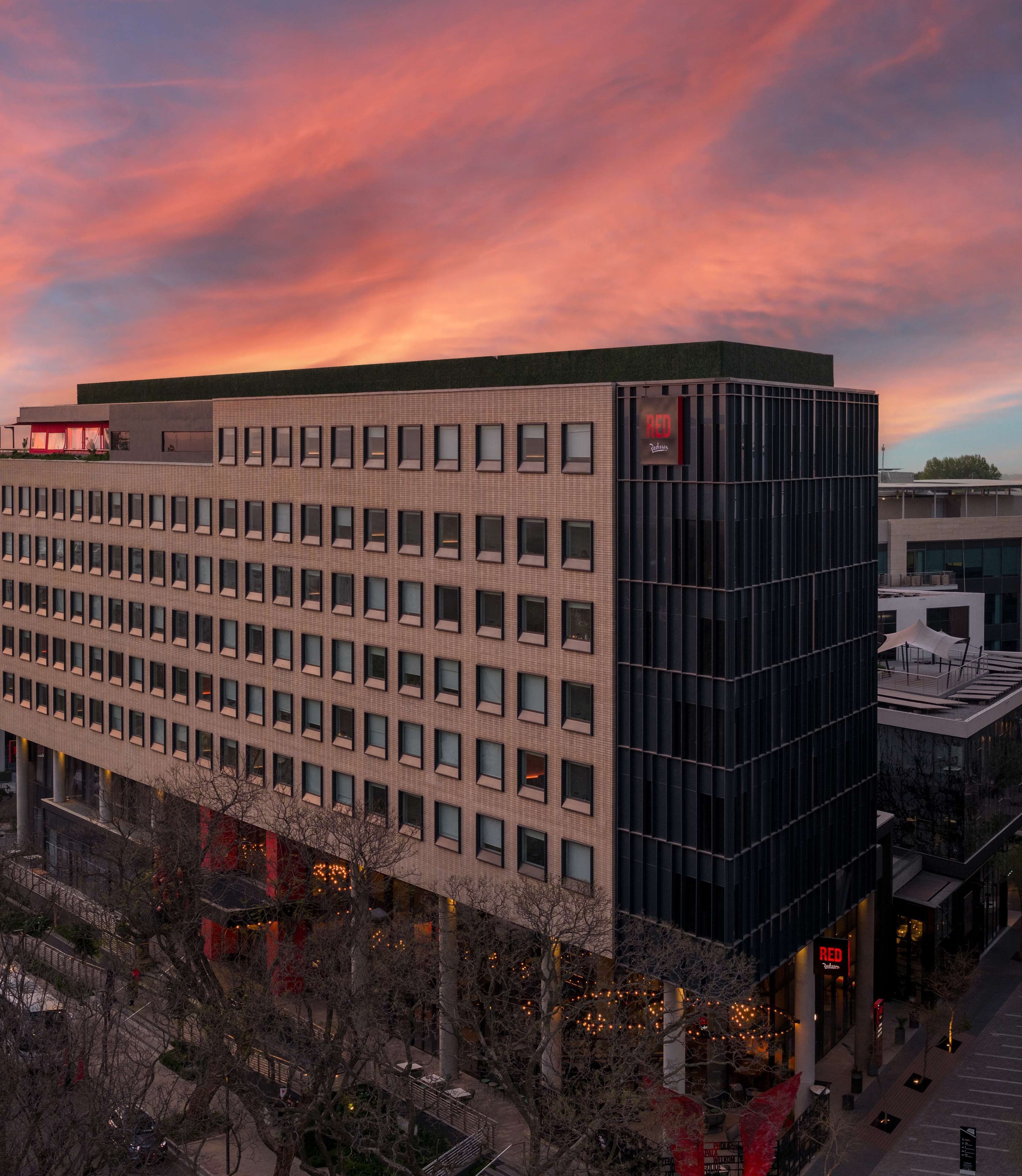 Un edificio moderno con una fachada de ventanas en forma de cuadrícula, un llamativo letrero que dice 'RED' y vista del cielo al atardecer.
