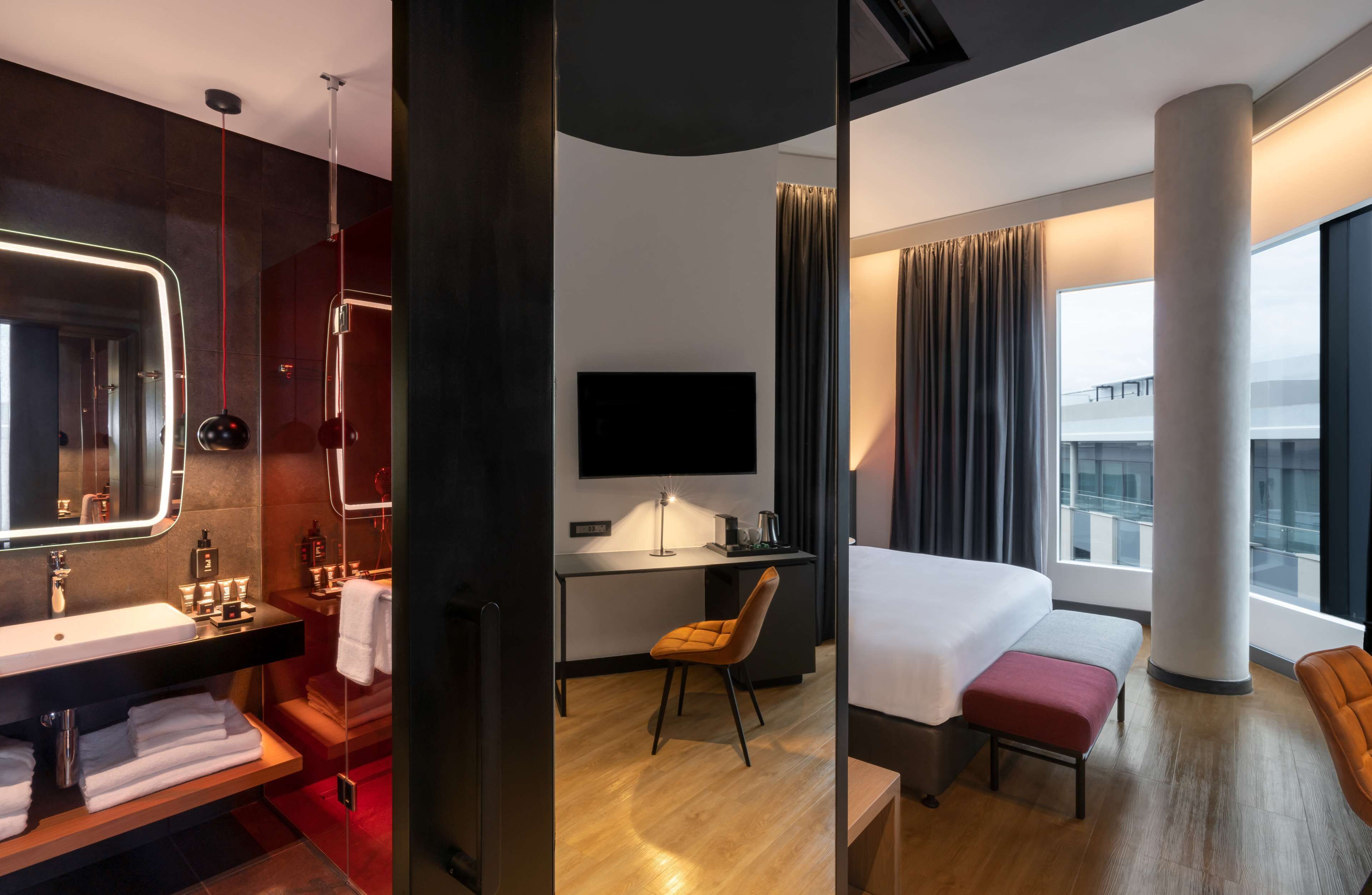 A modern hotel room with a large bed, a desk with a flat-screen TV, a bathroom with a large mirror and a vanity, and a view of the city through large windows.