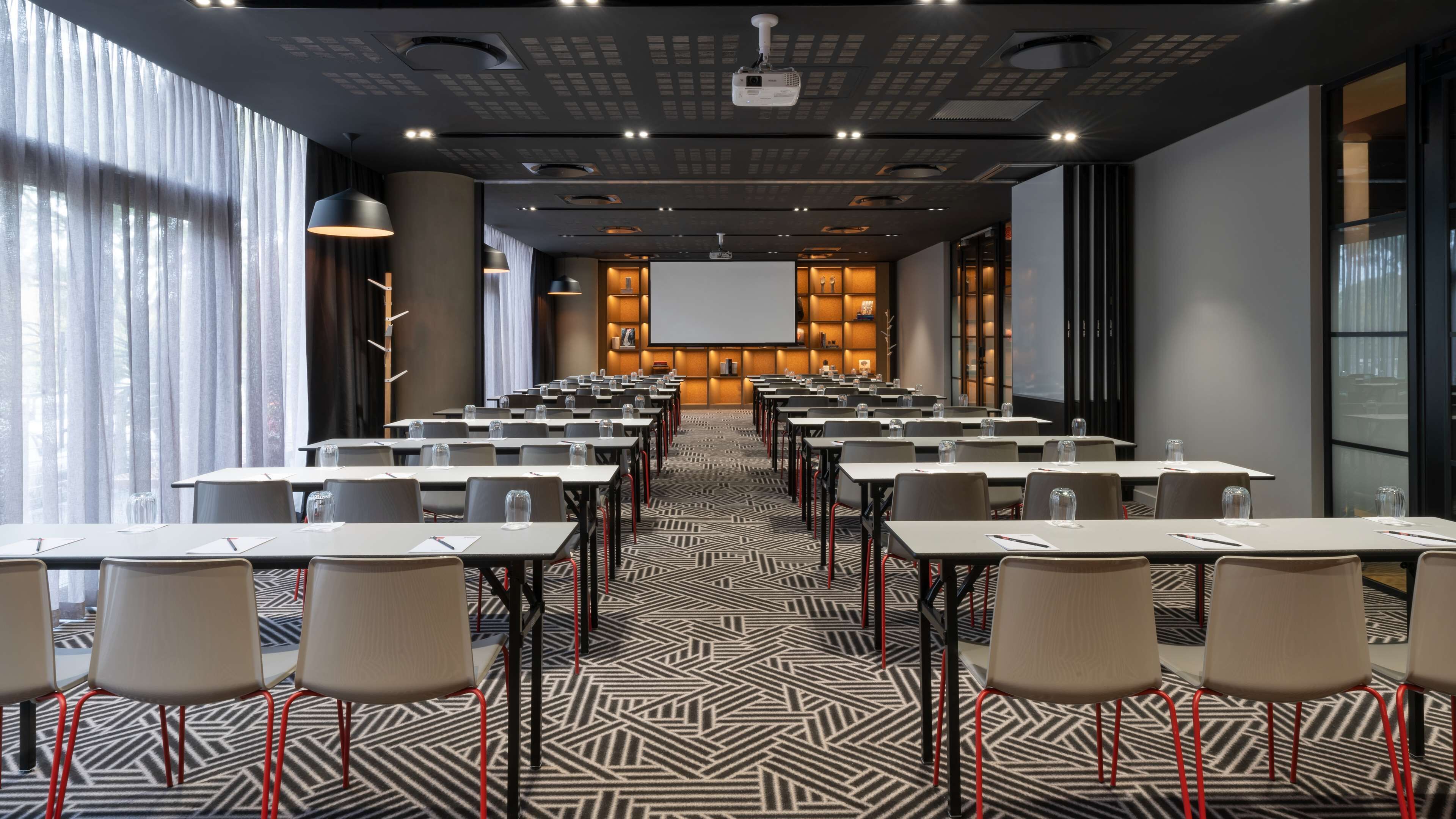 Un salón de conferencias con filas de mesas y sillas, una pantalla grande al frente y una alfombra estampada.
