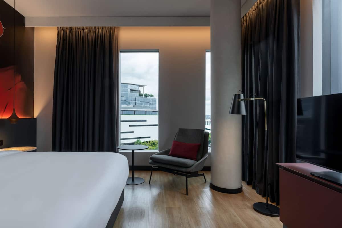 Habitación de hotel moderna con una cama grande, una mesita pequeña, una silla, un televisor y una ventana con vistas a un edificio.