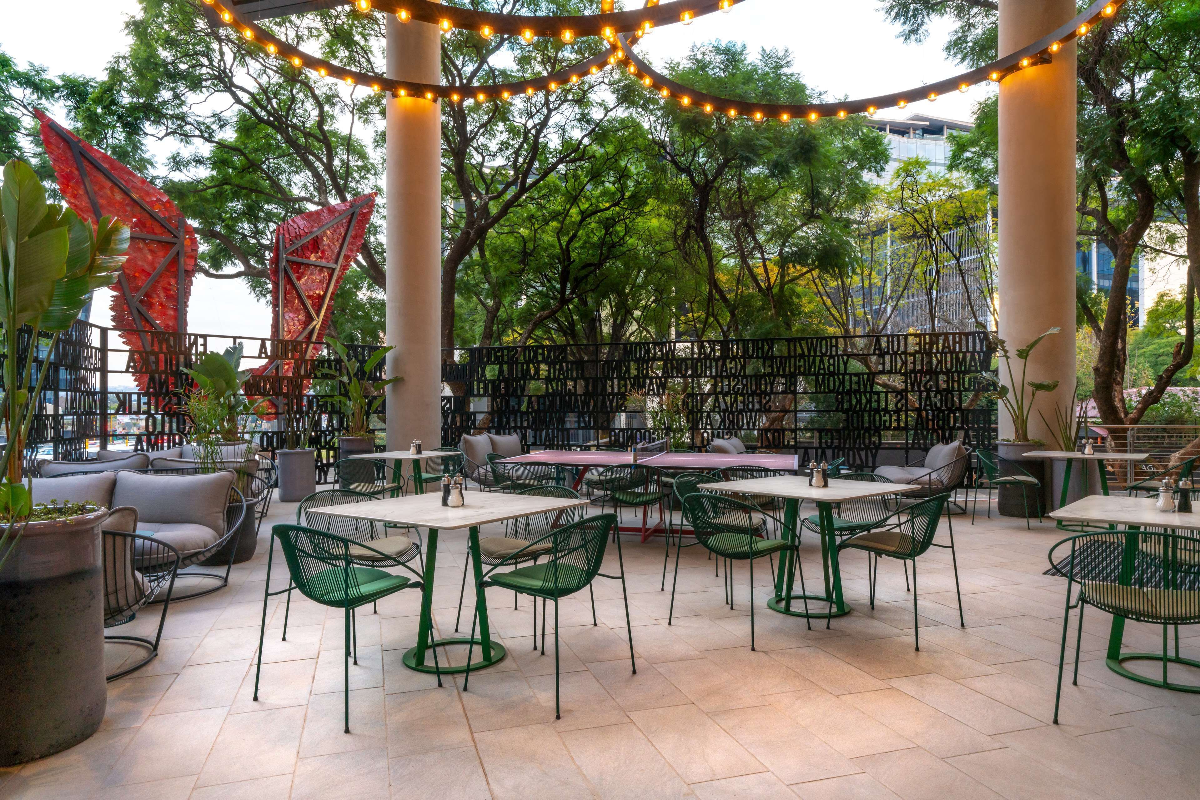 An outdoor cafe with tables and chairs, surrounded by trees and a metal fence.