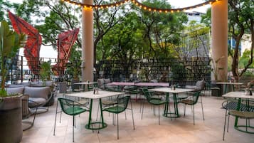 An outdoor cafe with tables and chairs, surrounded by trees and a metal fence.
