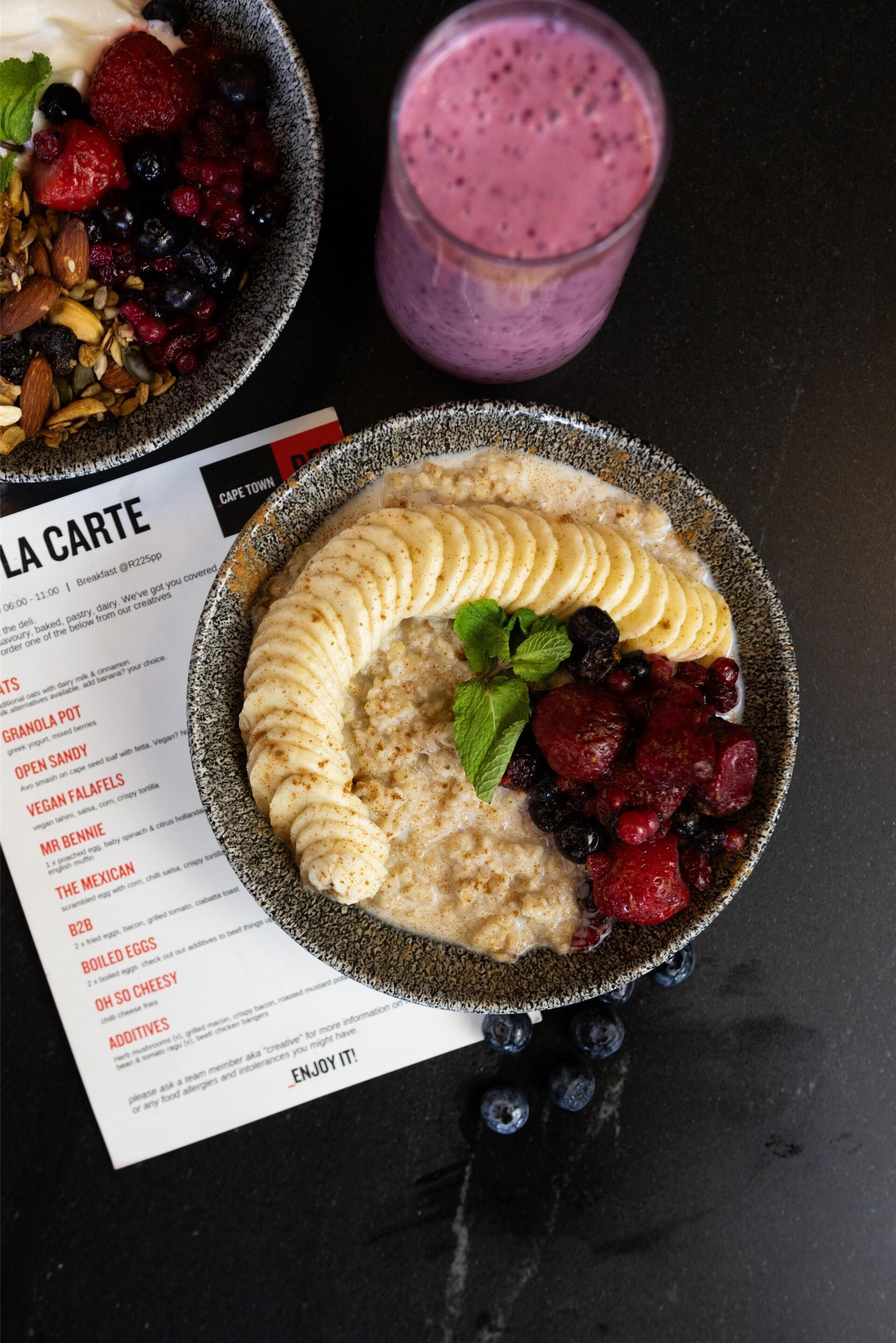 Una colazione con una ciotola di porridge ricoperta di banane, frutti di bosco e noci, un bicchiere di frullato e un menu.