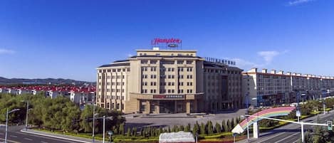 A large, multi-story building with a prominent 'Hampton' sign on top, surrounded by a landscaped area with trees and a pedestrian walkway.